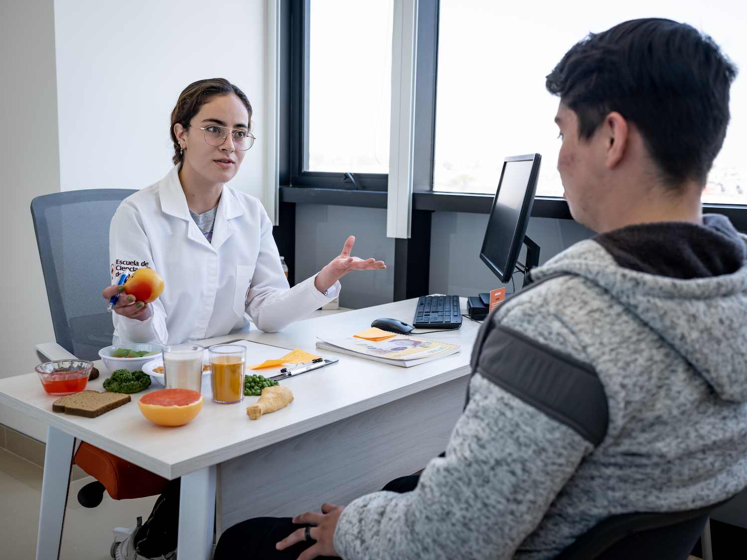 Nutrióloga en consulta explica hábitos alimenticios a un paciente, mostrando una manzana y alimentos saludables sobre el escritorio.